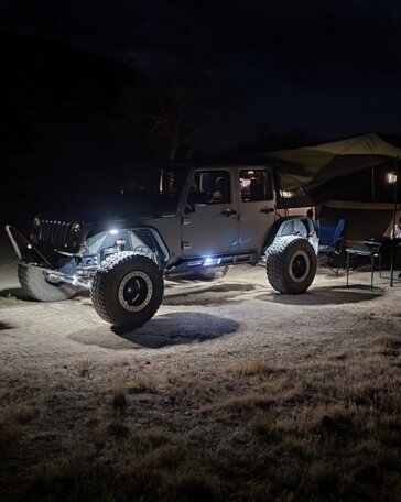 Jeep lit up by rock lights at a night camp, tent and chairs glowing in the background