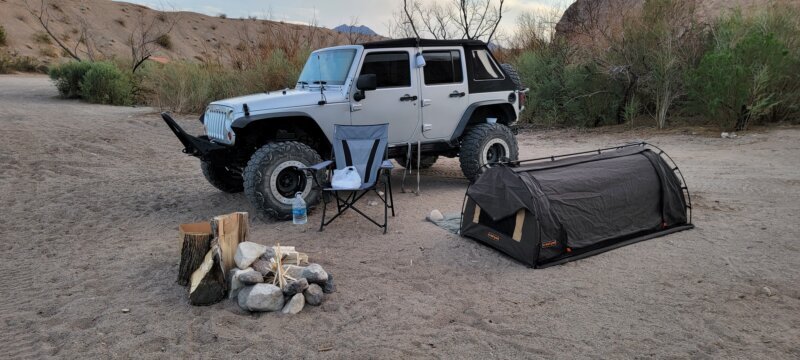 Rock-lined fire ring with stacked firewood at a desert camp, Jeep Wrangler and swag tent in background