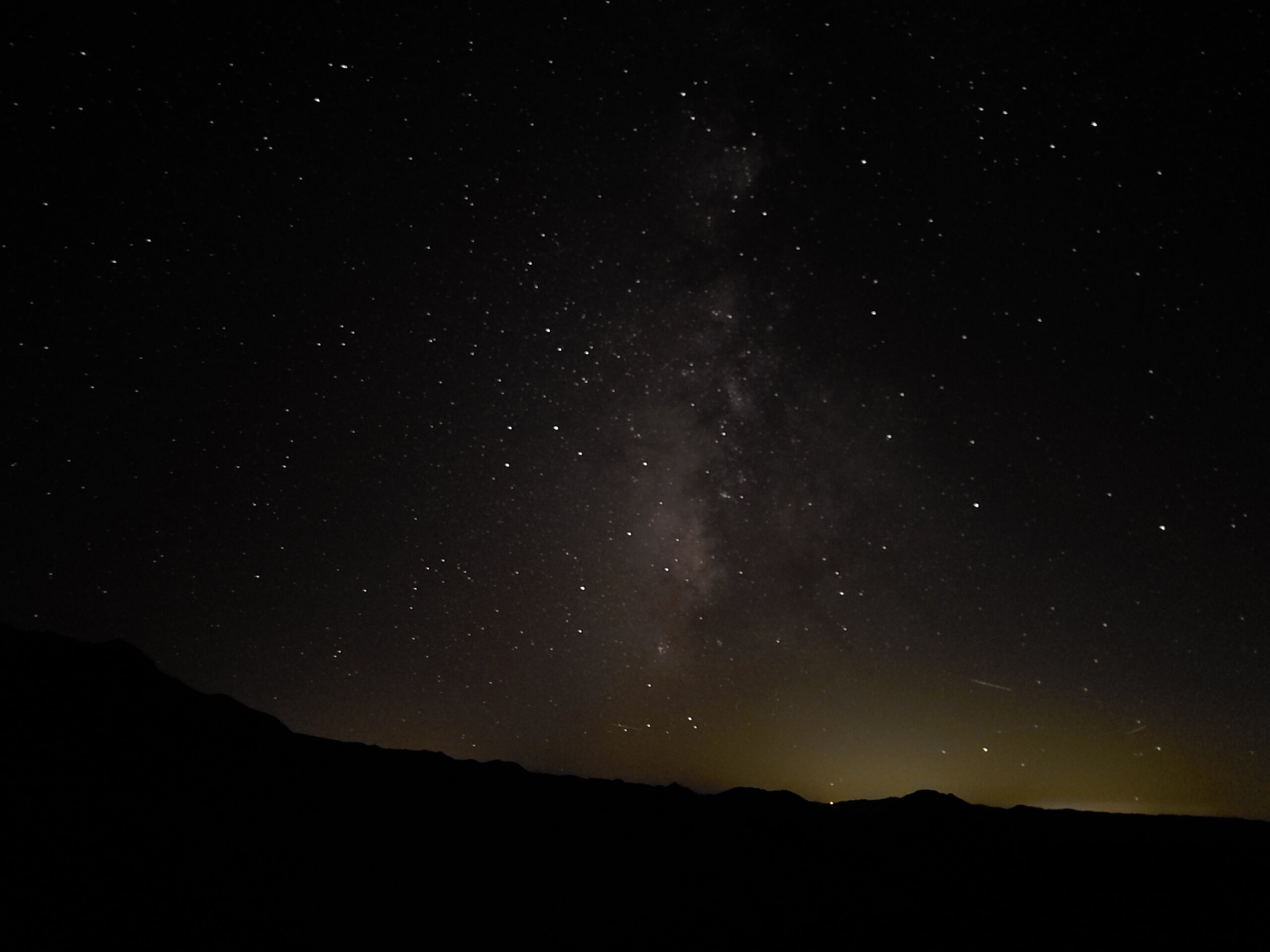Unedited phone night mode photo of the Milky Way band over a dark desert mountain ridge, with a faint glow of distant town light pollution on the horizon